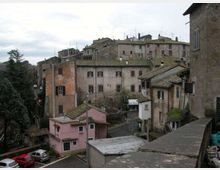 Veduta di un borgo collinare con edifici in muratura addossati tra loro e tetti in tegole, affacciati su una strada stretta. In primo piano si vedono un muretto e piccole case, mentre sullo sfondo compaiono alberi e un cielo nuvoloso.