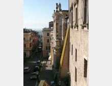 Vista dall’alto di una strada urbana in discesa affiancata da edifici storici in pietra e facciate ornate con balconi e cornici. In fondo si intravede l’orizzonte sul mare, con auto parcheggiate lungo i lati della via.
