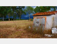 Piccolo edificio rurale intonacato con tetto in tegole rosse, con una porta decorata appoggiata all’esterno, ai margini di un campo erboso. Sullo sfondo si vedono alberi alti e un cielo scuro e nuvoloso.