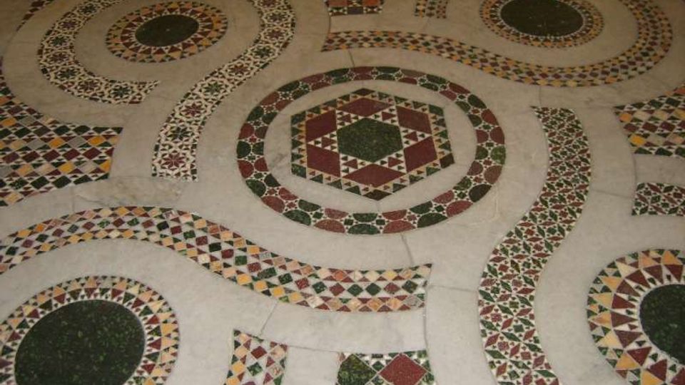 Pavimento decorativo in mosaico con grandi motivi circolari e ottagonali intrecciati, realizzati con tessere geometriche multicolori. La superficie chiara è intervallata da fasce e rosoni con pattern ripetuti in verde, rosso, giallo e nero.