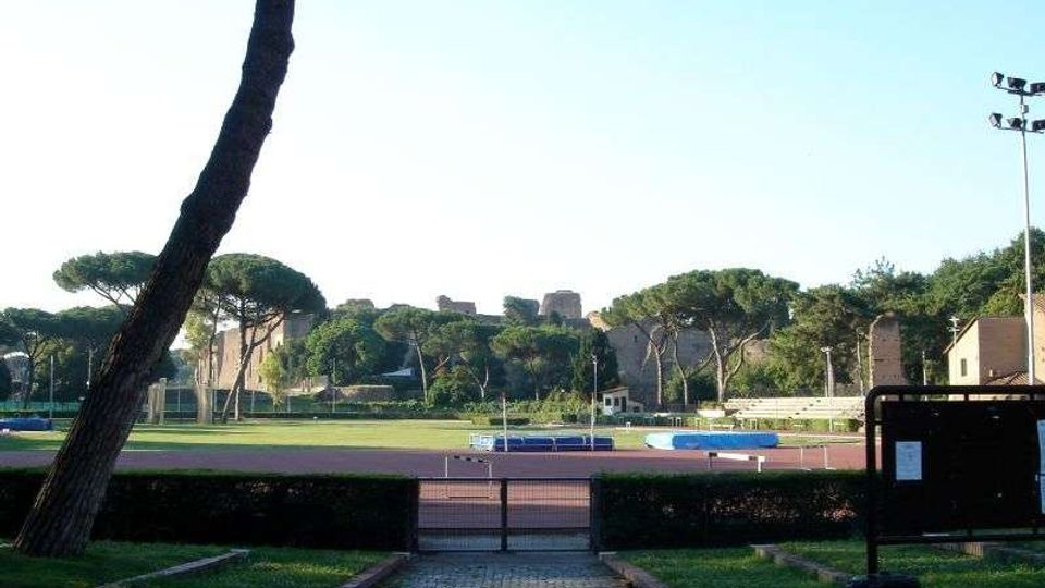 Vista di un impianto sportivo all’aperto con pista di atletica e campo centrale, circondato da siepi e alberi ad alto fusto. Sullo sfondo si vedono antiche mura in pietra e un’area verde con strutture e illuminazione da stadio.