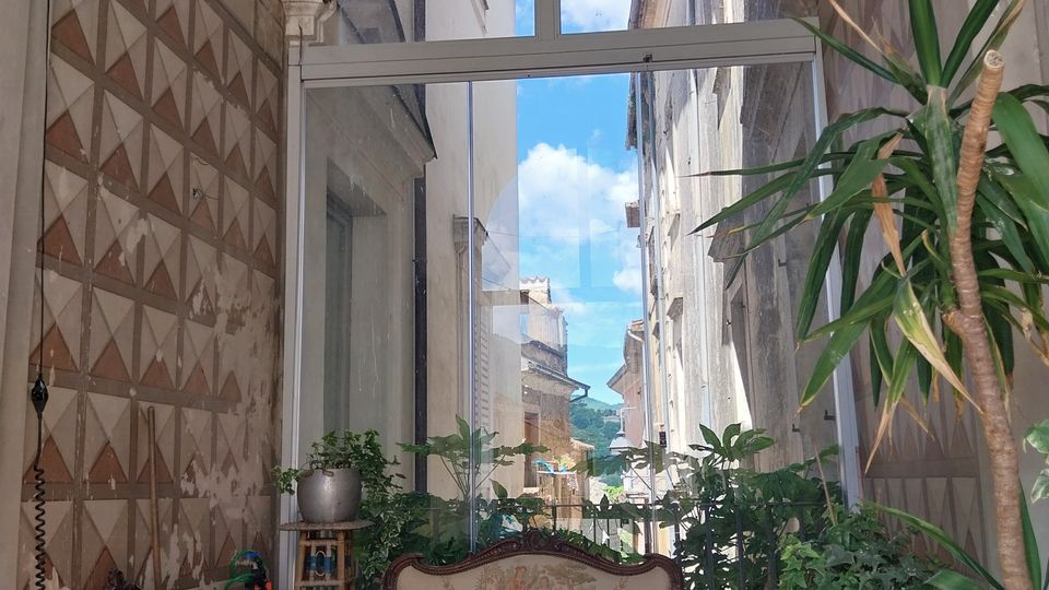 Piccolo atrio coperto con volta decorata e pareti a motivi geometrici, arredato con divanetto e sedie in stile classico tra molte piante. In fondo una grande vetrata ad arco lascia vedere una stretta via tra edifici in pietra sotto il cielo.