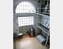 Interno di un loft a doppia altezza con grandi finestre ad arco e rettangolari a griglia che illuminano un ampio spazio. Sulla destra ci sono una struttura soppalcata in metallo con scala e parapetti, lampade a sospensione e arredi essenziali su pavimento piastrellato.