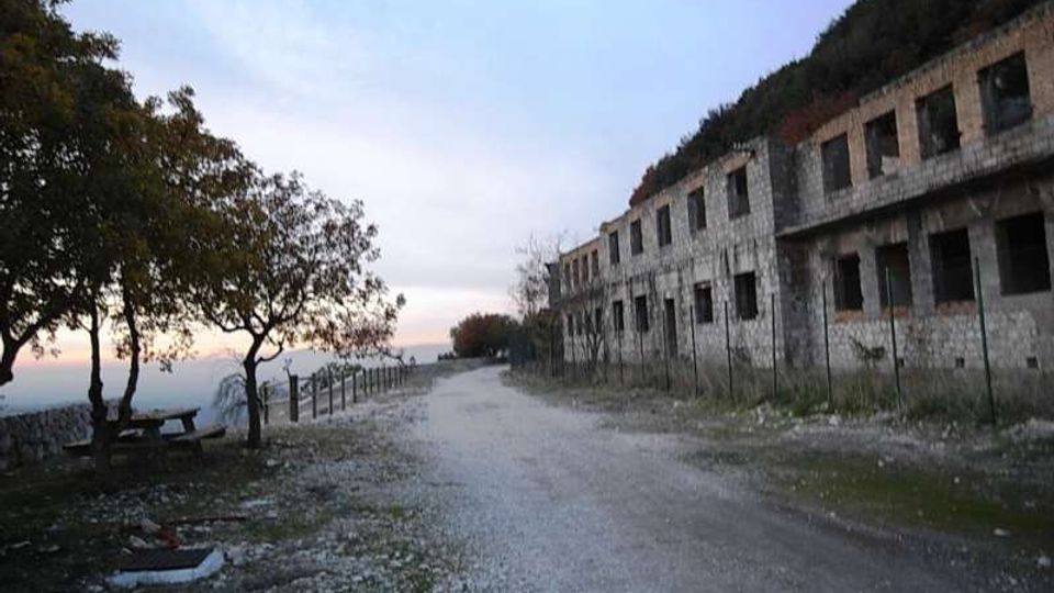 Strada sterrata lungo una scogliera, con alberi e panchine sul lato sinistro e un lungo edificio in pietra abbandonato con finestre vuote sul lato destro. Sullo sfondo si intravede il mare e l’orizzonte al tramonto.
