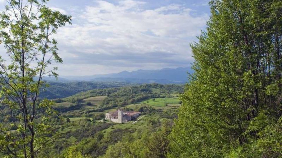 Vista panoramica su una valle collinare verde con campi e boschi, con un complesso edilizio in pietra su un crinale al centro. Alberi in primo piano incorniciano il paesaggio, con montagne lontane sotto un cielo parzialmente nuvoloso.