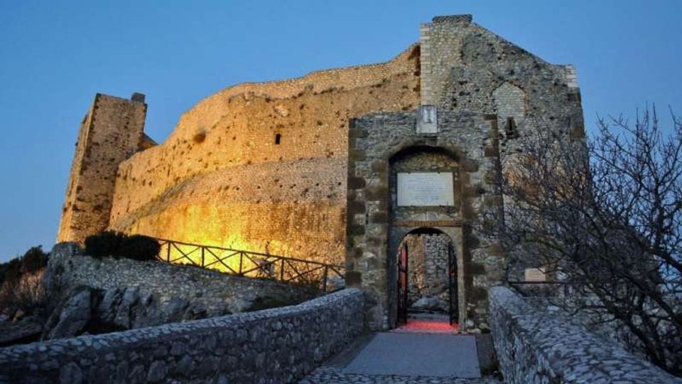 Veduta di un castello medievale in pietra su un’altura, con alte mura illuminate al crepuscolo. In primo piano si vede l’ingresso ad arco con portale e una passerella in pietra delimitata da muretti e ringhiere.