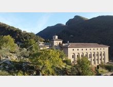 Complesso monastico in pietra su un pendio collinare, con lunga facciata a due piani scandita da arcate e un edificio centrale con campanile. Sullo sfondo si vedono montagne boscose e vegetazione mediterranea intorno alla struttura.