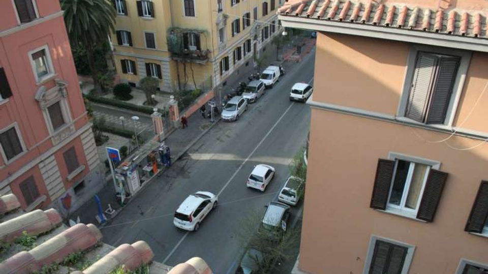 Vista dall’alto di una strada urbana con auto parcheggiate e in transito, fiancheggiata da edifici residenziali dai colori pastello con persiane e tetti in tegole. Sul lato sinistro si vede un piccolo giardino recintato con palme e un’area d’ingresso lungo il marciapiede.