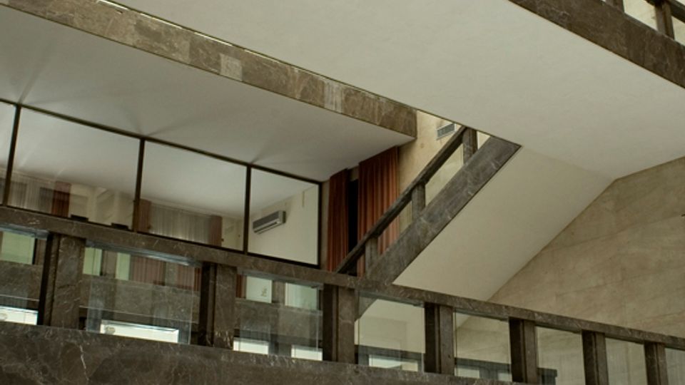Atrio interno di un edificio moderno a più livelli, con passerelle e rampe in pietra scura e parapetti in vetro. Un lucernario a griglia sul soffitto illumina lo spazio con grandi superfici vetrate e un’area centrale con panchine.
