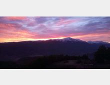 Panorama montano al tramonto con una lunga cresta in controluce e una cima parzialmente innevata sullo sfondo. Il cielo è attraversato da nuvole rosa e arancioni, con colline scure e vegetazione in primo piano.