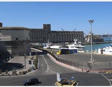 Veduta di un porto industriale con banchine e imbarcazioni ormeggiate, affiancato da grandi edifici in cemento tipo silos e magazzini. In primo piano si vedono una strada curvilinea con incrocio, recinzioni e lampioni lungo l’area portuale.