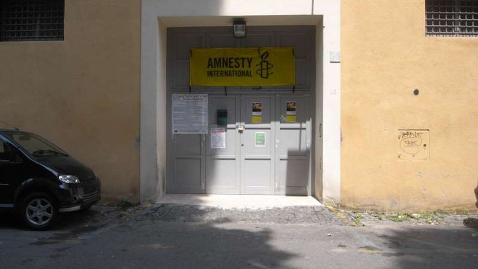 Ingresso di un edificio con grande portone grigio incassato in una facciata color ocra, con un’insegna gialla “Amnesty International” sopra la porta e alcuni avvisi affissi. Davanti c’è un’area asfaltata con ombre marcate e una finestra con grata visibile sulla parete.