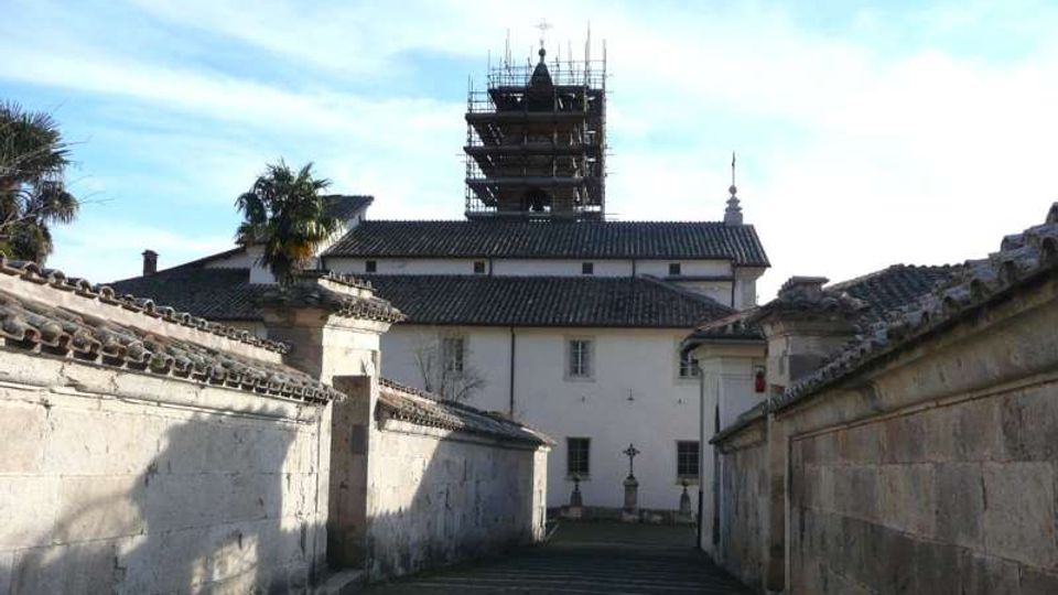 Vicolo in discesa fiancheggiato da alti muri intonacati con tetti in coppi, che conduce a un edificio bianco in stile tradizionale. Sullo sfondo si vede una torre con impalcature di restauro e una piccola cupola, sotto un cielo azzurro.