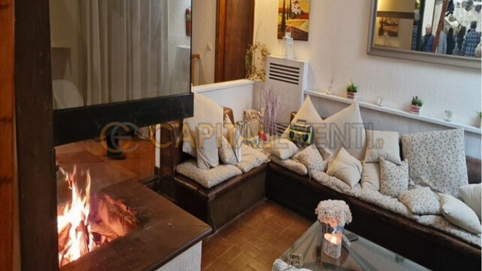 Salotto interno con caminetto a legna acceso in muratura e divano ad angolo con numerosi cuscini. Tavolino in vetro con centrino, pareti chiare con mensole e decorazioni, e specchio a parete che riflette parte dell’ambiente.