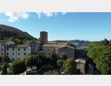 Complesso di edifici in pietra e intonaco con tetti in tegole e una torre campanaria quadrata al centro, circondato da alberi. Sullo sfondo si vedono colline verdi e un cielo azzurro con alcune nuvole.