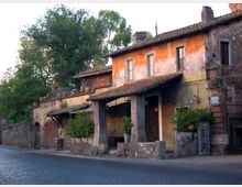 Casa rurale in pietra e intonaco color ocra, con tetto in tegole e piccolo portico su pilastri, affacciata su una strada asfaltata. Sul lato si vedono un cancello, vasi e muretti bassi, con alberi alti sullo sfondo.