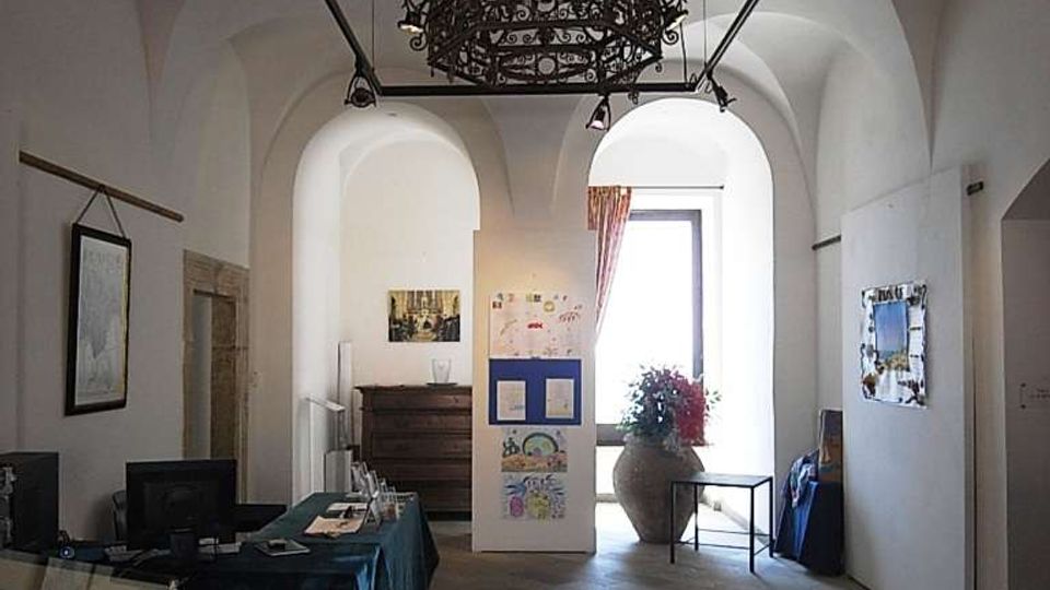 Interno di una sala espositiva con soffitto a volte bianche e grande lampadario in ferro battuto sospeso. Pavimento in pietra grigia, scrivania con computer a sinistra e pannelli espositivi con una grande finestra luminosa sul fondo.