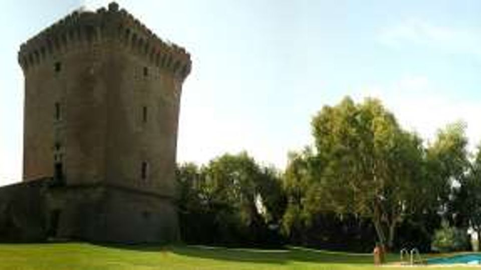 Torre in pietra medievale a pianta quadrata su un terrapieno, circondata da un ampio prato verde e da alberi. In primo piano si vede una lunga piscina rettangolare con bordo azzurro sotto un cielo sereno.