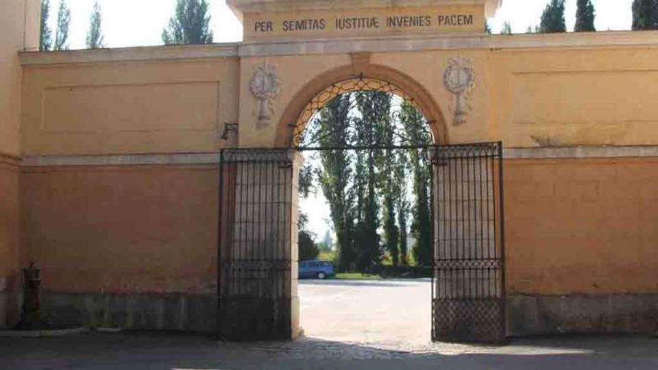 Ingresso monumentale con arco centrale in muratura color ocra, sormontato da una scritta latina e decorazioni in rilievo. Un grande cancello in ferro battuto aperto conduce a un viale con alberi alti sullo sfondo.