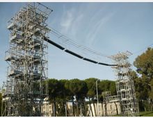 Due alte torri metalliche a traliccio con piattaforme e scale interne sostengono una passerella sospesa collegata da cavi. La struttura si trova all’aperto, con alberi sullo sfondo e edifici bassi nelle vicinanze sotto un cielo sereno.
