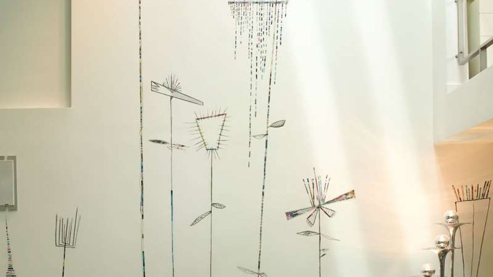 Soggiorno moderno a doppia altezza con parete bianca decorata da grandi disegni stilizzati di fiori verticali e divani modulari verde lime. A destra si vede un soppalco con scaffali e ringhiera, illuminato dalla luce naturale dall’alto, con lampade da terra in metallo.