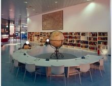 Interno di una biblioteca moderna con grande tavolo circolare bianco e sedie, al centro un mappamondo antico su supporto metallico. Lungo la parete ci sono scaffali bassi pieni di libri e una grande mappa appesa, con ampie vetrate sul lato sinistro.