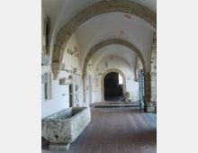 Corridoio interno con volte a botte e grandi archi in pietra su pareti intonacate bianche, pavimento in mattoni a spina di pesce e oggetti lapidei esposti lungo le pareti. In primo piano si vede una vasca in pietra, mentre sul fondo un arco conduce a un ambiente più scuro con una finestra.