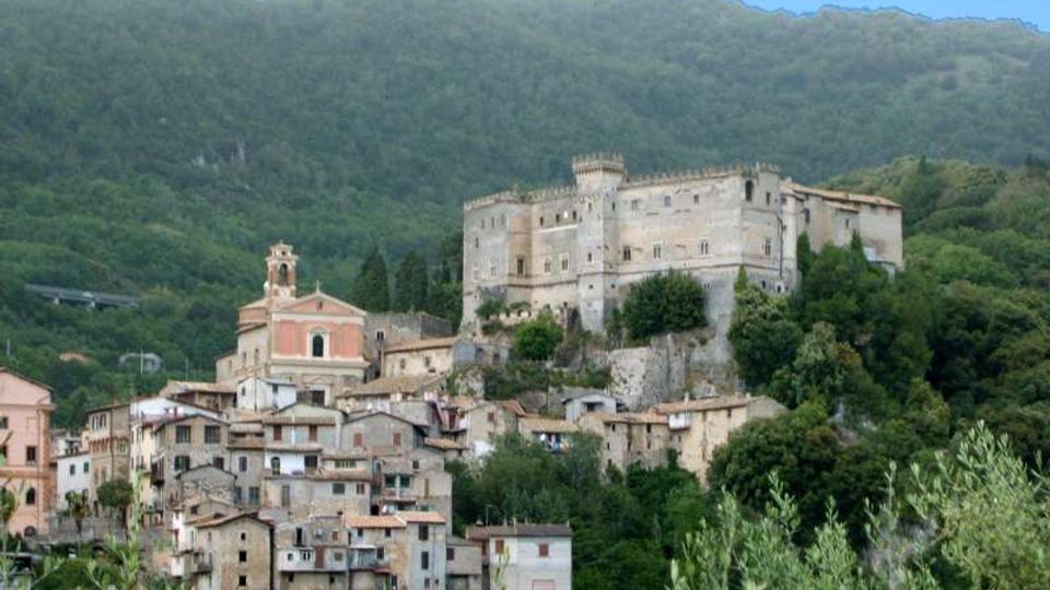 Veduta di un borgo collinare con case in pietra e una chiesa con campanile, dominato da un grande castello fortificato su uno sperone roccioso. Sullo sfondo si estendono pendii montuosi ricoperti di boschi.