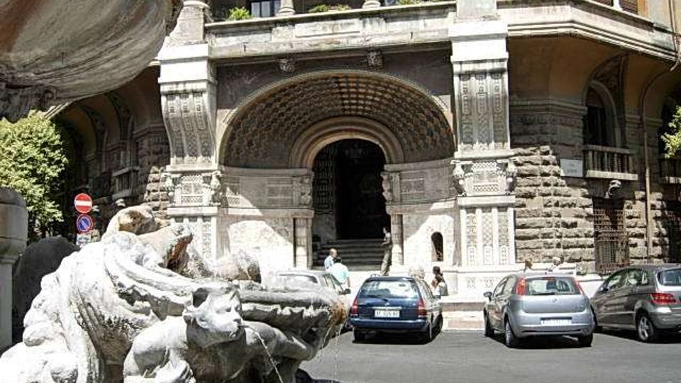 Ingresso di un grande edificio storico in pietra con arco monumentale decorato e colonne, affacciato su una strada urbana. In primo piano si vede una fontana in marmo con figure scolpite e vasca, con auto parcheggiate davanti all’entrata.
