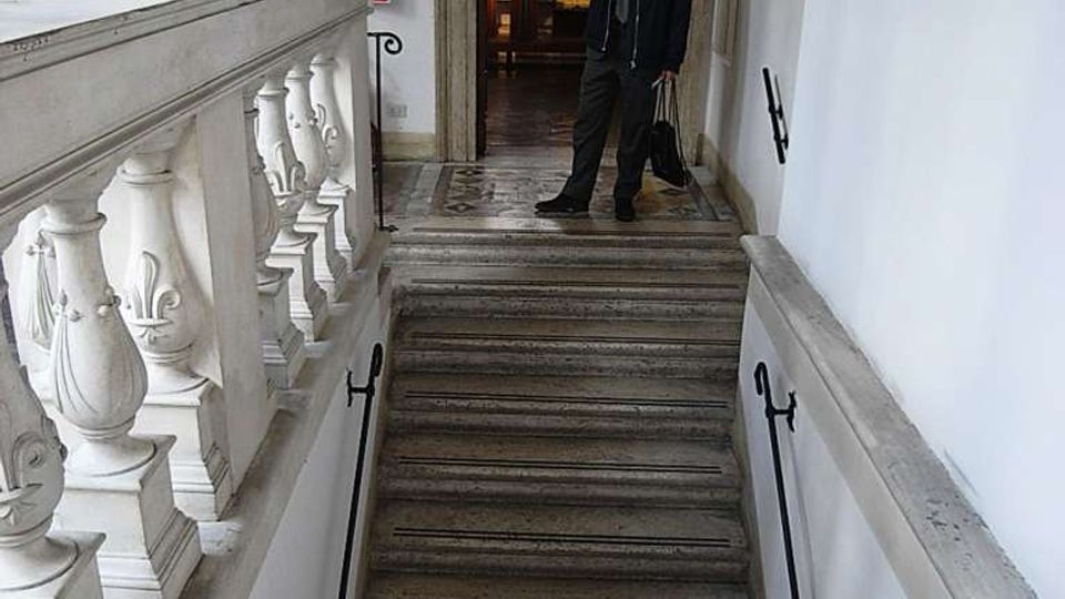 Scala interna in pietra con gradini larghi, corrimano in metallo e balaustra in marmo decorata. In cima alle scale si vede un pianerottolo con pavimento a mosaico e una porta con cornice in pietra che conduce a una stanza illuminata da una finestra.