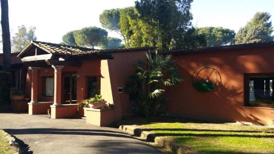 Edificio basso con facciata in intonaco color terracotta, ingresso con piccolo portico a colonne e tetto in tegole, affacciato su un vialetto asfaltato curvo. Intorno ci sono prato e alberi alti, con una finestra e una decorazione circolare sulla parete.