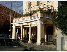 Edificio ad angolo in stile classico con portico a colonne e balconata superiore, con ringhiera e fioriere su una terrazza. Davanti si vede un marciapiede in pietra con arredi esterni e piante in vaso, affacciato su una strada urbana con lampione e auto parcheggiate.