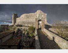 Antica fortificazione in pietra su uno sperone roccioso, con alte mura e una torre laterale sotto un cielo nuvoloso. Un ponte rialzato lastricato conduce a un portale ad arco d’ingresso, affiancato da bassi edifici con tetti in tegole.
