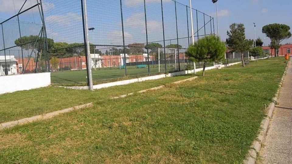 Campo sportivo recintato con alte reti metalliche e pali per l’illuminazione, affiancato da un prato con piccoli alberi. Sullo sfondo si vedono edifici bassi e una strada pedonale lungo il bordo destro.