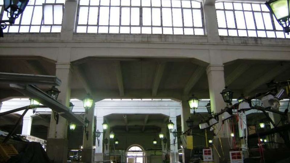 Interno di un grande edificio industriale o mercato coperto con alte colonne e travi in cemento, ampie finestre superiori a griglia e lampioni sospesi. Lungo i lati si vedono banchi e strutture metalliche sotto una copertura alta con luce naturale che entra dalle finestre.