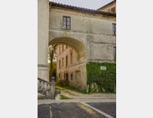 Passaggio ad arco sotto un edificio storico in muratura intonacata, con tetto in coppi e finestre a riquadri, che conduce a un cortile interno. In primo piano si vede una strada acciottolata con una breve scalinata in pietra, e sulla parete a destra un tratto coperto di edera con una piccola targa.