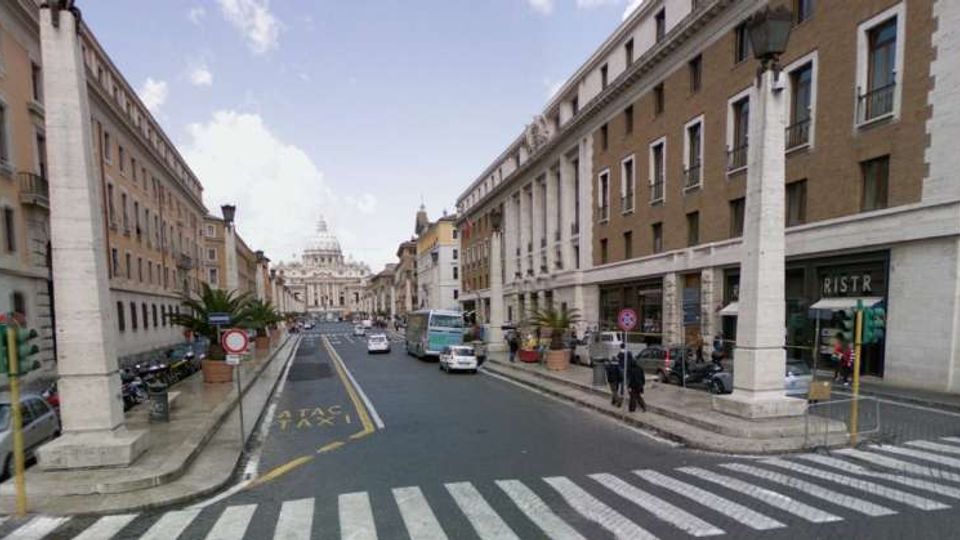 Strada urbana ampia con attraversamento pedonale in primo piano, fiancheggiata da alti edifici in muratura e porticati, con grandi pilastri bianchi e lampioni ai lati. In lontananza si vede la cupola e la facciata della Basilica di San Pietro, con veicoli e segnaletica stradale lungo la carreggiata.