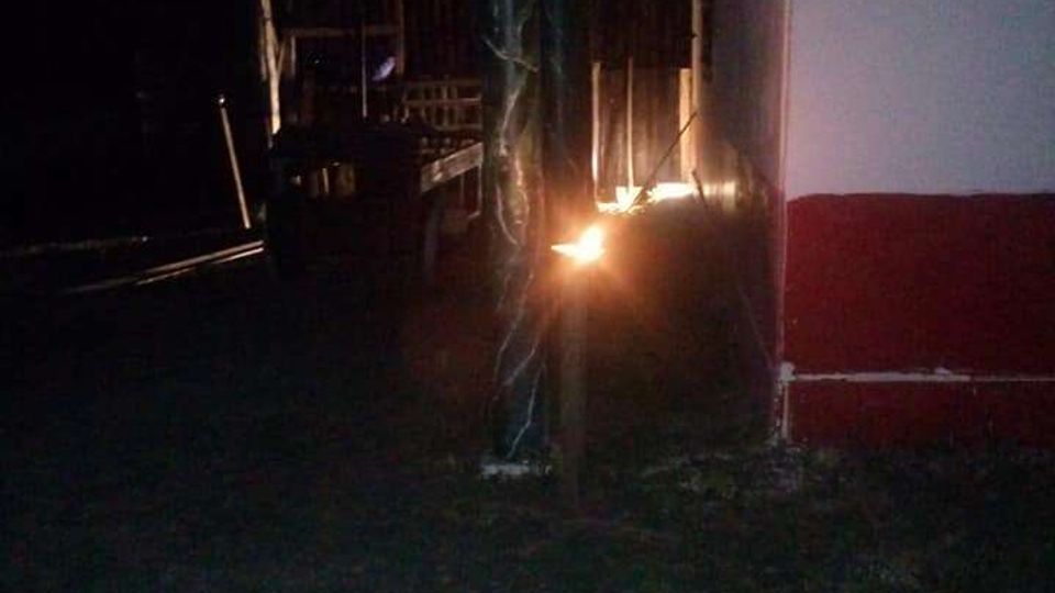 Veranda esterna di notte con tetto in legno sorretto da pali, affiancata a un edificio bianco con fascia rossa alla base. In primo piano c’è un prato e sotto la tettoia si vedono arredi e una piccola fonte di luce vicino a uno dei pilastri.