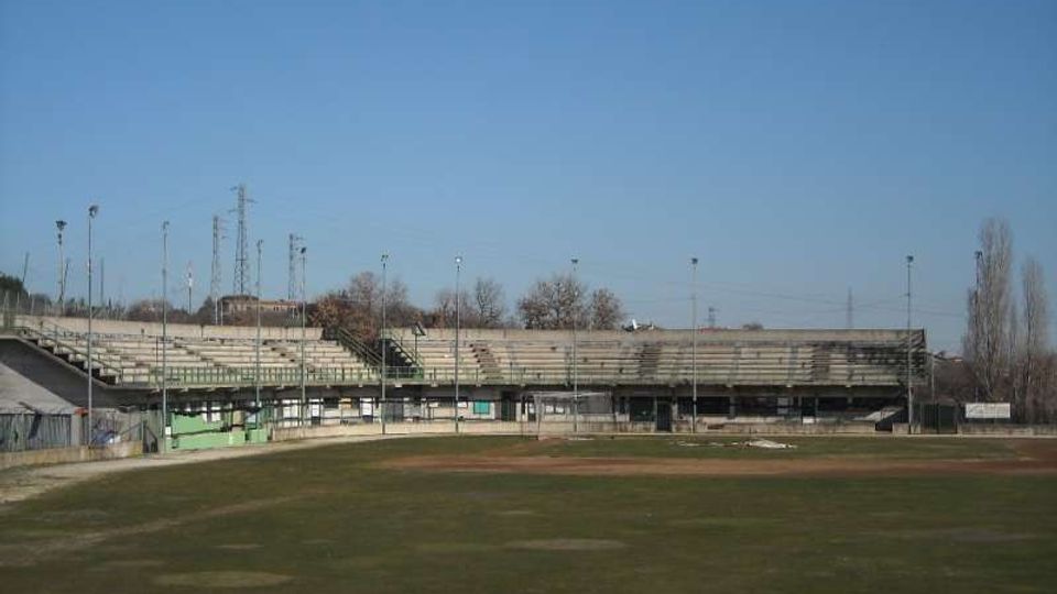 Campo sportivo all’aperto con ampia area in erba e pista in terra battuta, circondato da tribune in cemento con gradinate e pali per l’illuminazione. Sullo sfondo si vedono alberi spogli e infrastrutture elettriche sotto un cielo sereno.