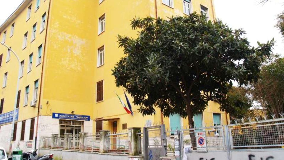 Edificio scolastico a più piani con facciata gialla e finestre con persiane verdi, circondato da recinzione metallica e muro con cartelli e graffiti. Davanti all’ingresso si vedono un albero grande, un’area di accesso con cancelli e una bandiera italiana.