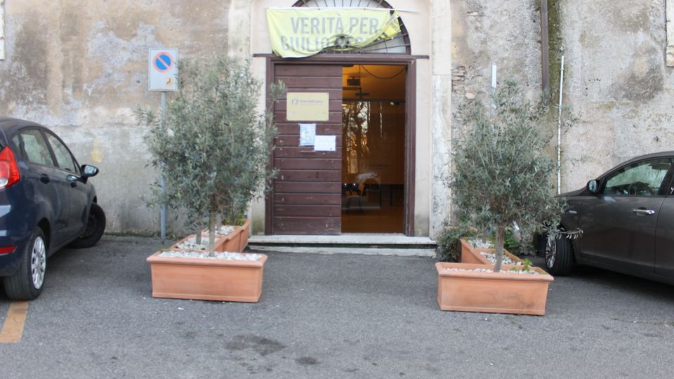 Ingresso di un edificio in muratura con intonaco consumato, portale ad arco e porta in legno aperta con luce interna visibile. Davanti all’entrata ci sono due grandi fioriere rettangolari con alberelli, affiancate da auto parcheggiate ai lati e un cartello stradale sulla parete.