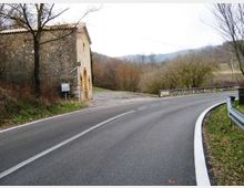 Strada asfaltata di campagna che curva a destra, con guardrail metallico e segnaletica a bordo carreggiata. A sinistra si vede un edificio in pietra vicino al margine della strada, con alberi spogli e colline sullo sfondo.