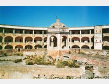 Cortile interno di una struttura storica in rovina, con un edificio semicircolare a due piani e una sequenza di arcate su entrambi i livelli. Al centro si vede un piccolo padiglione con cupola e portico, mentre il suolo del cortile è in parte spoglio e in parte invaso da vegetazione e detriti.