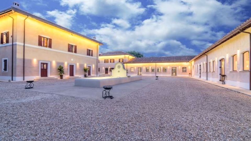 Cortile interno in ghiaia di un complesso edilizio con edifici bassi intonacati e tetti in tegole, illuminati da luci calde lungo le facciate. Al centro si trova una piccola fontana con muretto curvo, sotto un cielo parzialmente nuvoloso.