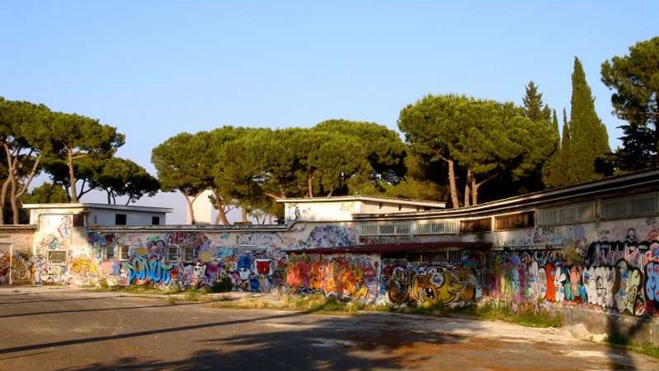 Ampio cortile asfaltato circondato da edifici bassi con tettoie e lunghe pareti ricoperte di graffiti colorati. Sullo sfondo si vedono grandi pini e cipressi sotto un cielo sereno.