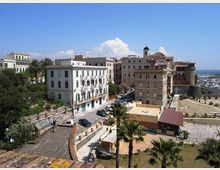 Veduta panoramica di un quartiere costiero con edifici storici e palazzi chiari, una strada con auto parcheggiate e palme in primo piano. Sullo sfondo si vede un porto turistico con numerose barche e il mare oltre le mura del lungomare.