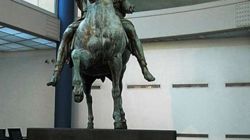 Atrio interno di un museo moderno con grandi superfici vetrate e pannelli luminosi sospesi sul soffitto. Al centro, su un basamento, una grande statua equestre in bronzo circondata da pareti chiare e una breve scalinata sullo sfondo.