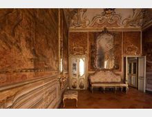 Interno di una sala storica in stile barocco/rococò con pareti affrescate e boiserie decorata, soffitto ornato e pavimento in legno. Al centro si vedono un grande specchio con cornice intagliata sopra un divano imbottito, una piccola panca e porte con pannelli decorativi.