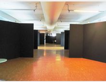 Ampio spazio interno tipo galleria espositiva con pavimento lucido e soffitto basso illuminato da luci lineari. Lungo il corridoio centrale sono presenti grandi pannelli divisori neri che formano aree separate.