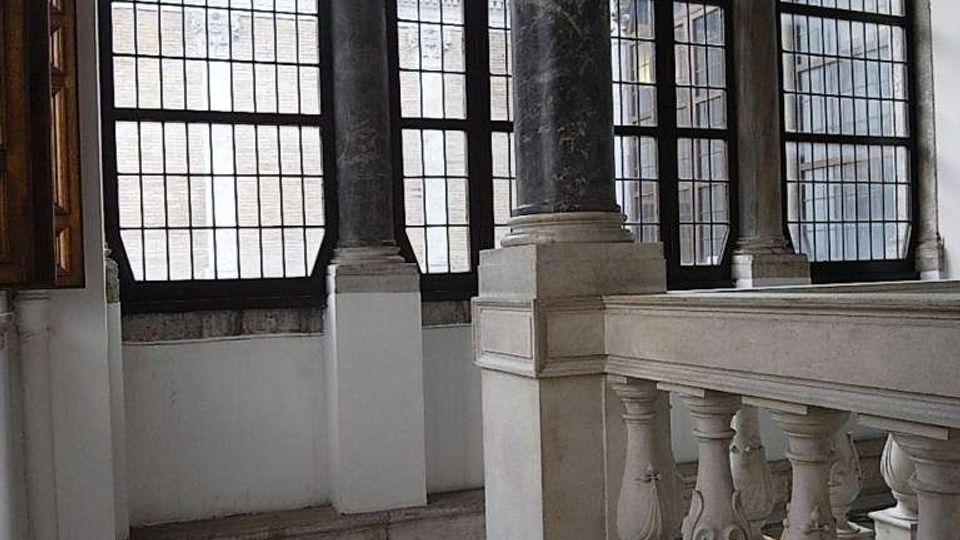 Interno di un edificio storico con ampie finestre a riquadri in ferro, colonne in pietra con capitelli decorati e pavimento a mosaico geometrico. In primo piano si vede una balaustra in marmo lungo una scala o un ballatoio.
